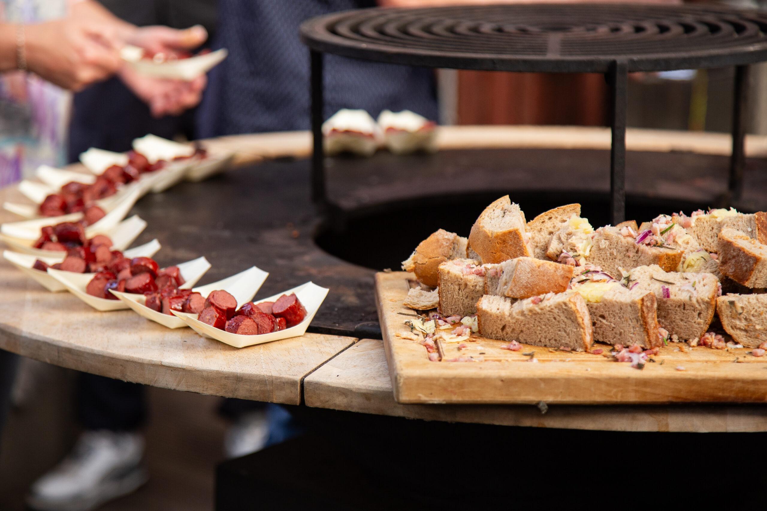 Hermann befüllt im BBQ Raum Hürth kleine Servierschiffchen auf dem Rand des Puncher-Grills mit Wurst und Sauce, im Vordergrund Brot mit Käse und Zwiebeln