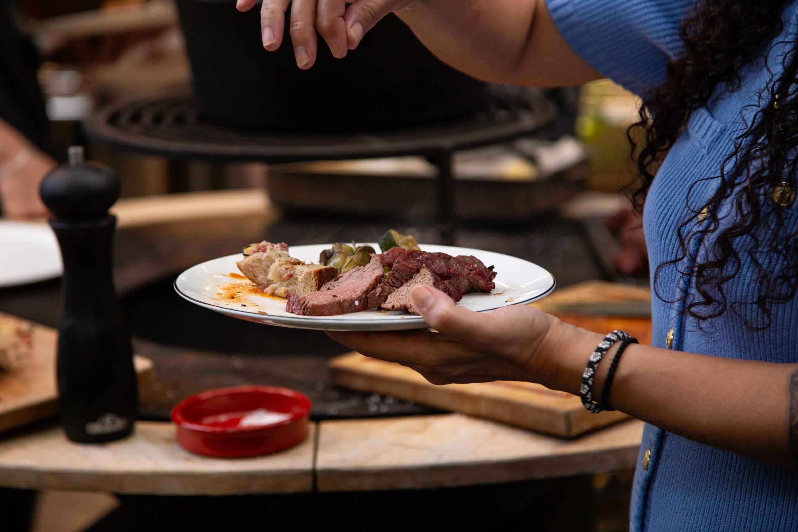 Eine Frau hält im BBQ Raum Hürth einen Teller mit frisch zubereitetem Fleisch in der Hand und streut grobes Salz darüber – im Hintergrund unscharf der Puncher-Grill
