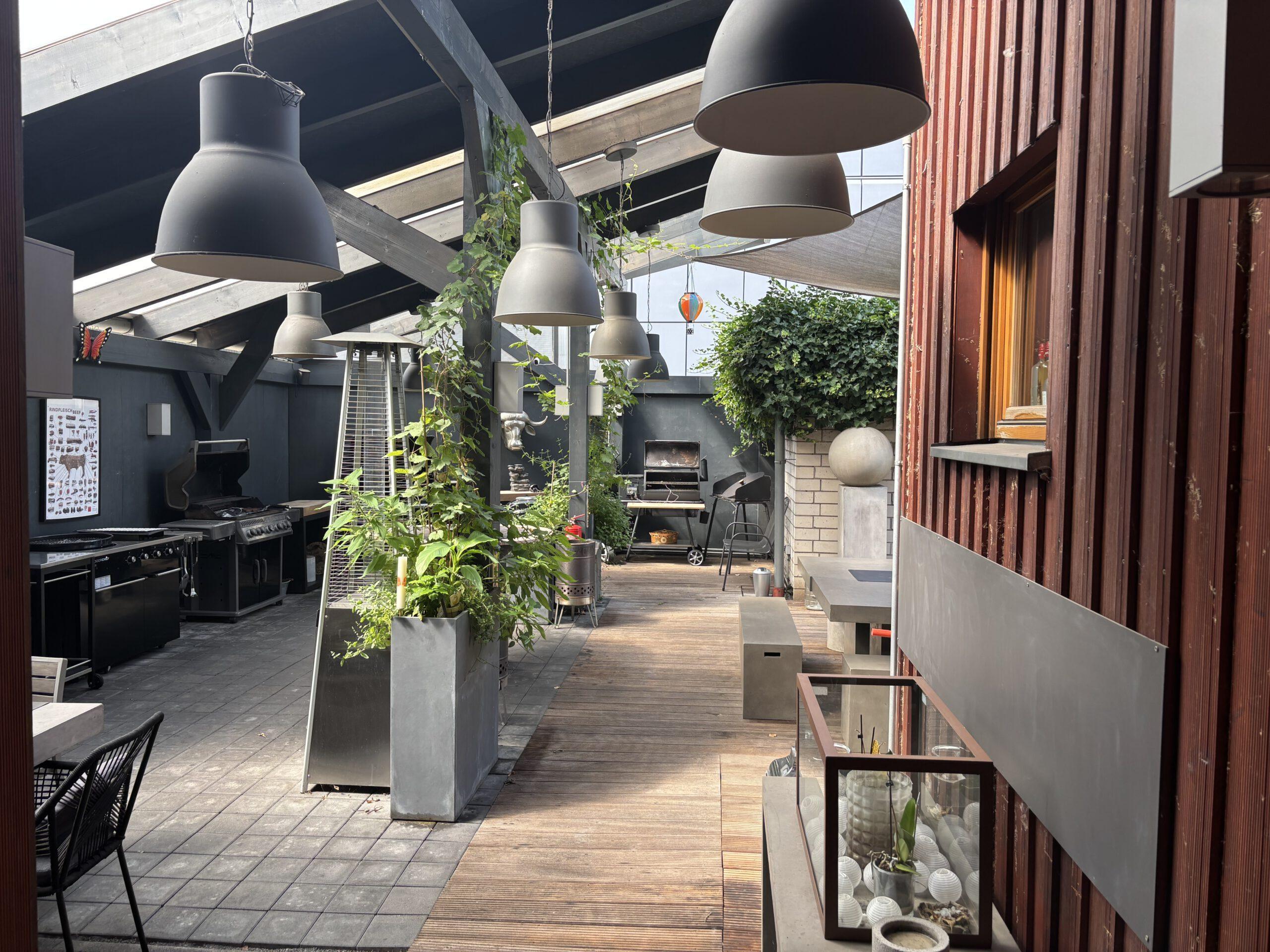 Blick von außen auf die Terrasse des BBQ Raum Hürth mit massivem Steintisch für 9 Personen, Überdachung, Stierkopf-Dekoration und Puncher im Hintergrund